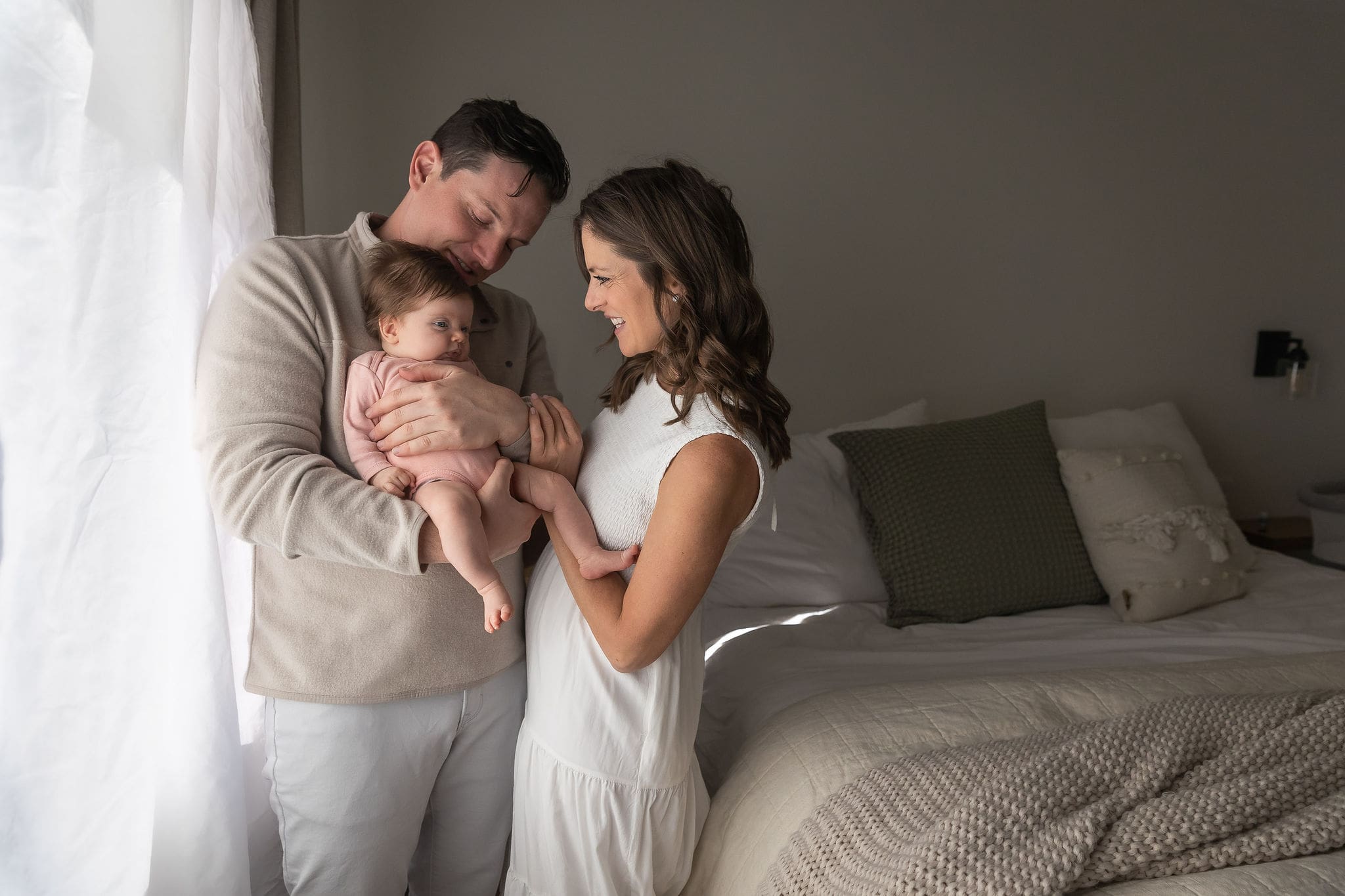 Happy new parents giggle with their newborn in dad's arms while standing in a window