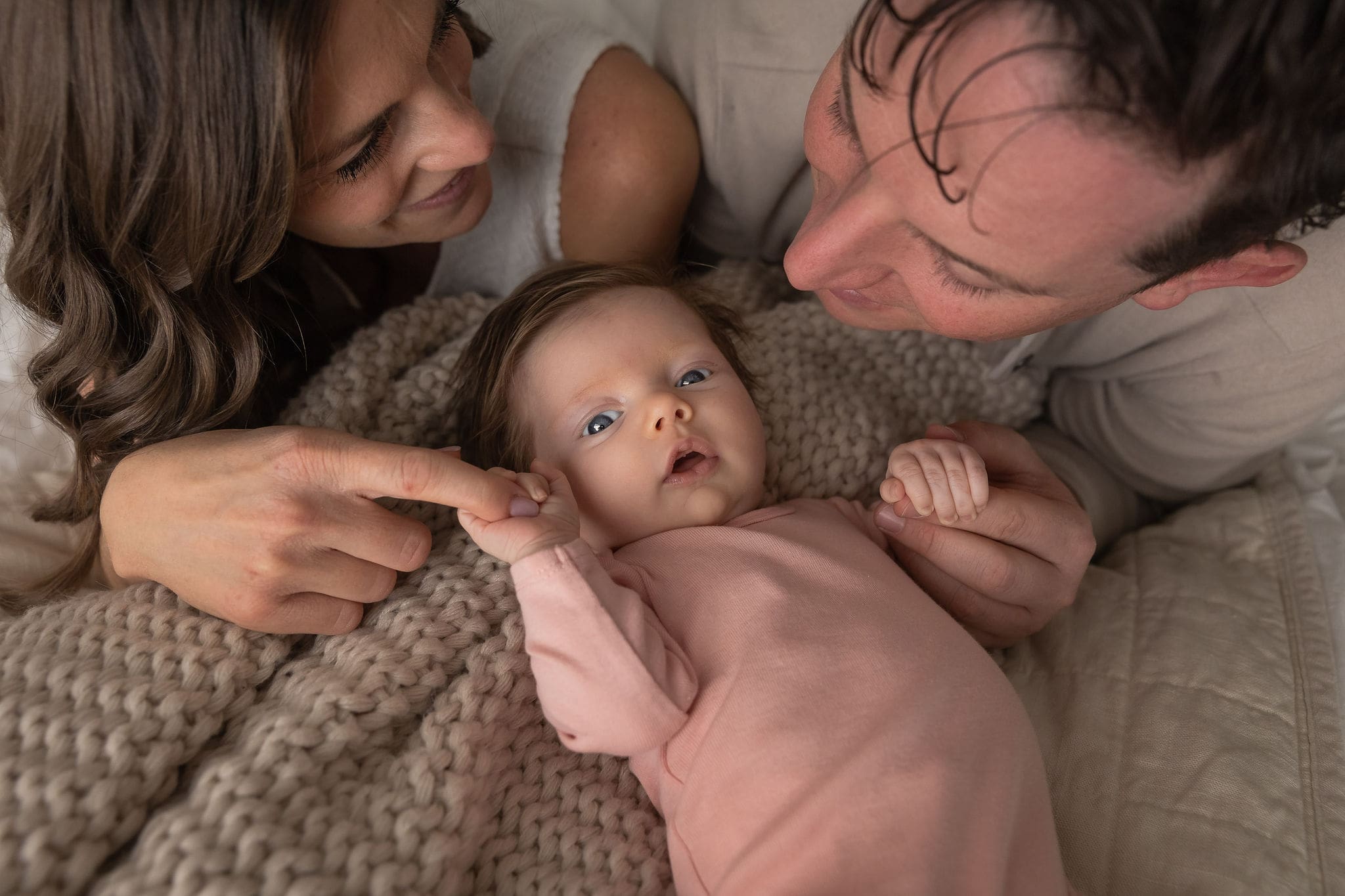 A newborn baby lays on a blanket holding mom and dad's fingers in a pink onesie after visiting baby boutiques in san diego