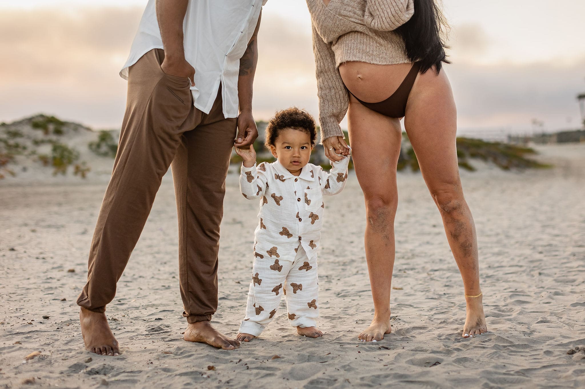 A toddler in teddy bear pajamas stands on a beach at sunset holding pregnant mom and dad's hands