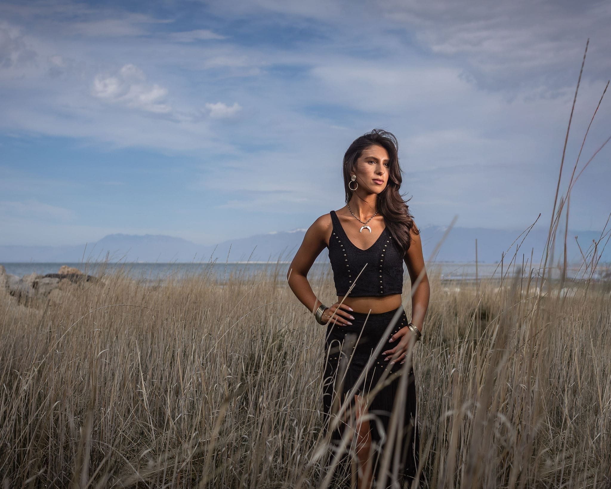 A woman in a black ensemble stands in tall golden grass on the beach with a hand on her hip