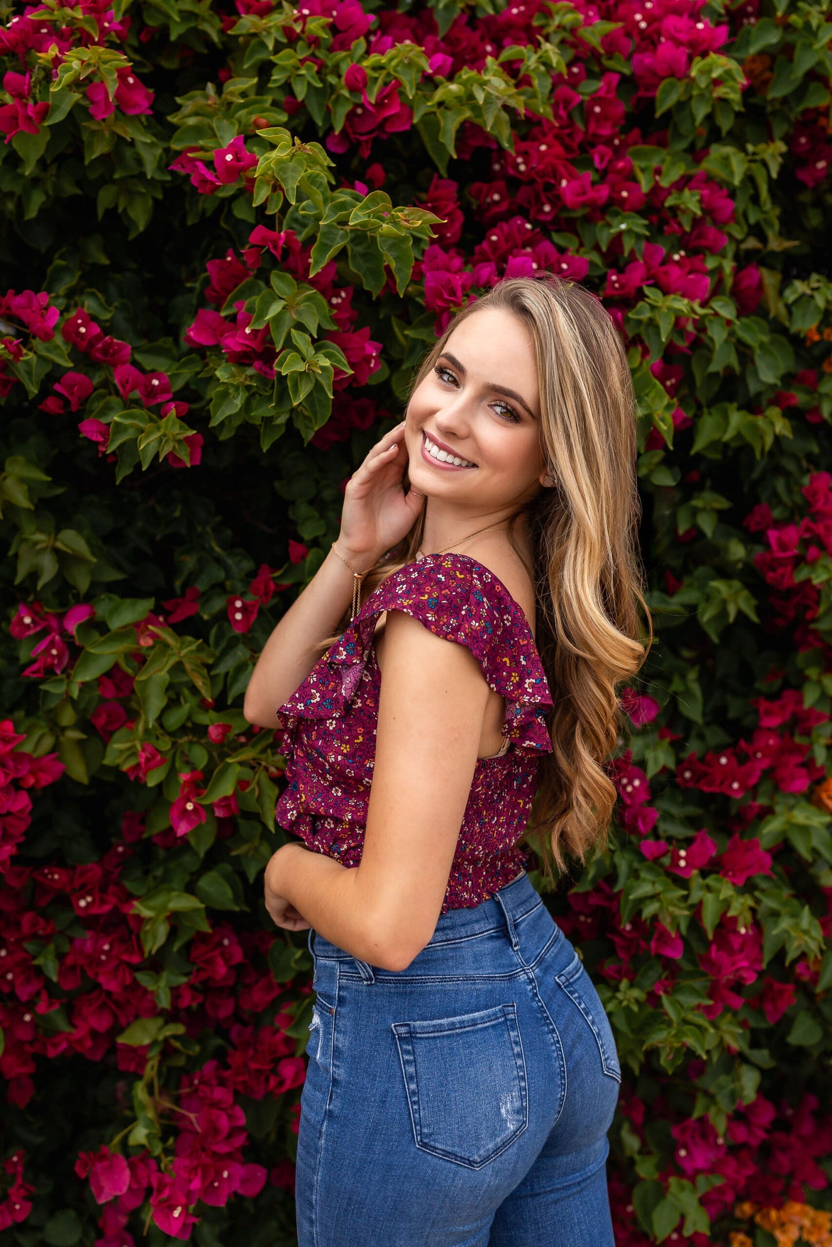 A smiling high school senior in a purple blouse and jeans stands against a flowering bush smiling over her shoulder after finding boutiques in san diego