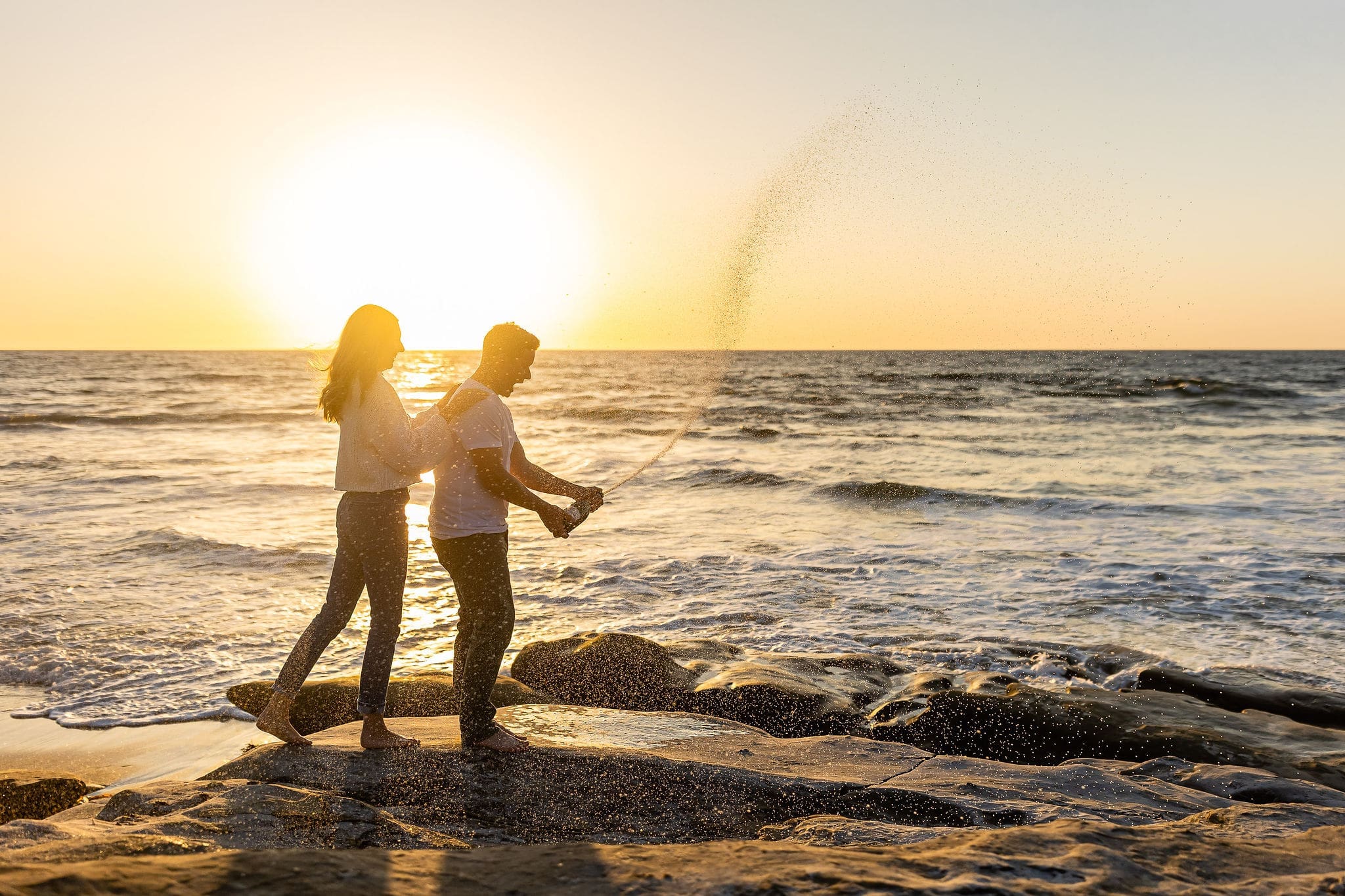 A mom sprays a bottle of champagne on a beach at sunset with his new fiancee