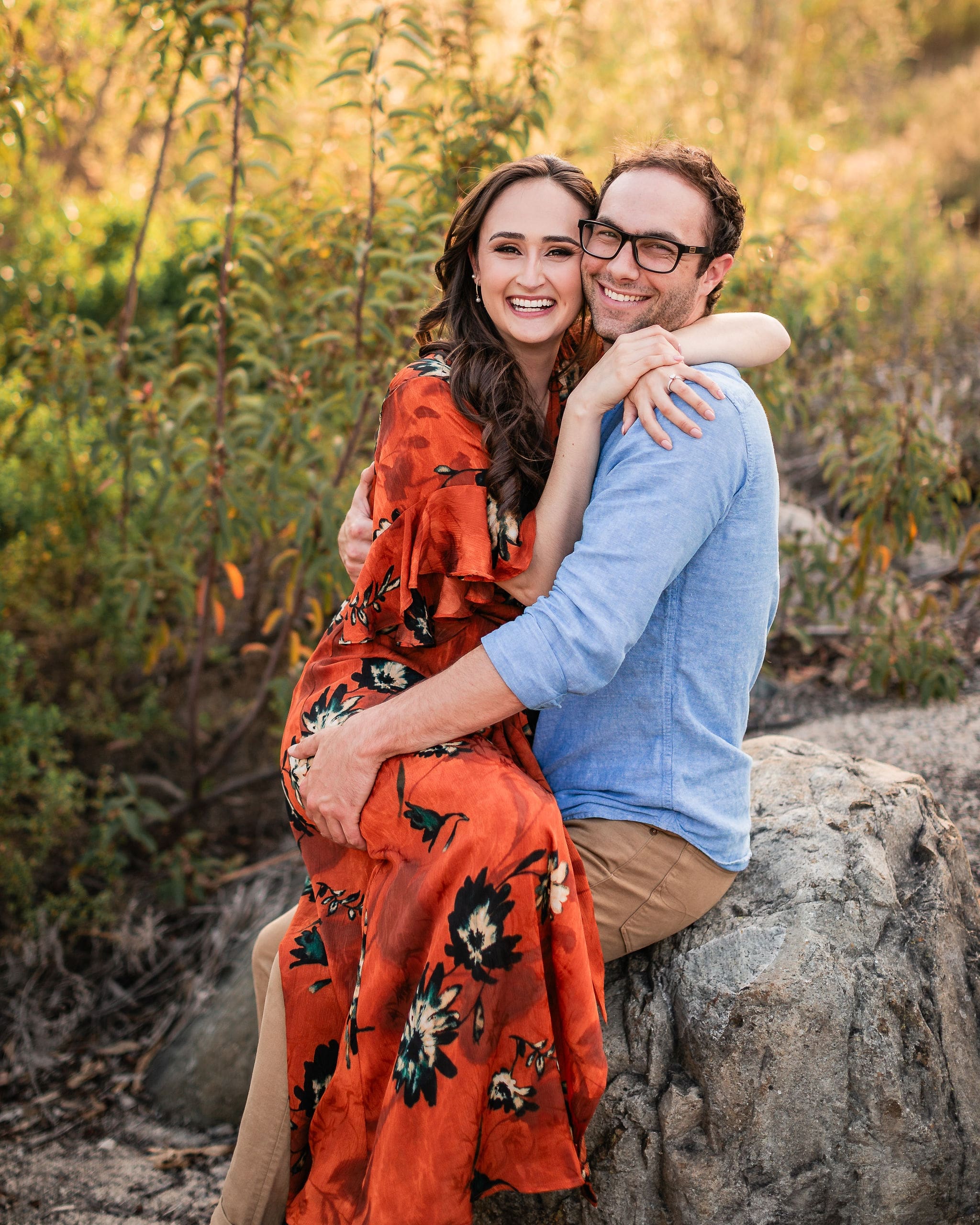 A happy engaged couple sits on a boulder smiling and hugging cheek to cheek at sunset in red and blue during one of the date night ideas in san diego