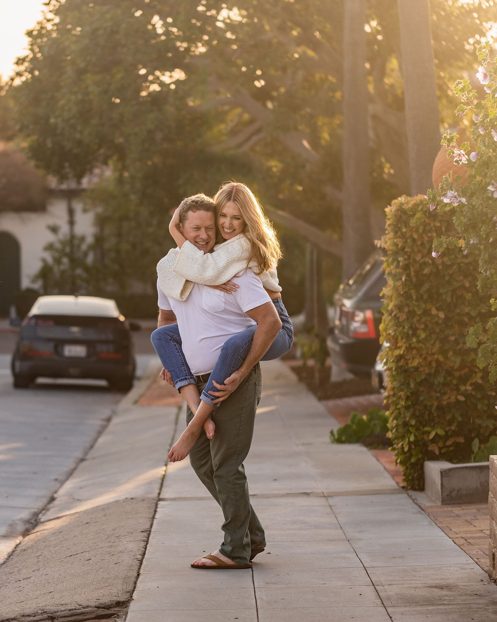 A happy woman in jeans is given a piggy back ride by her fiancee on a street sidewalk at sunset after finding date night ideas in san diego