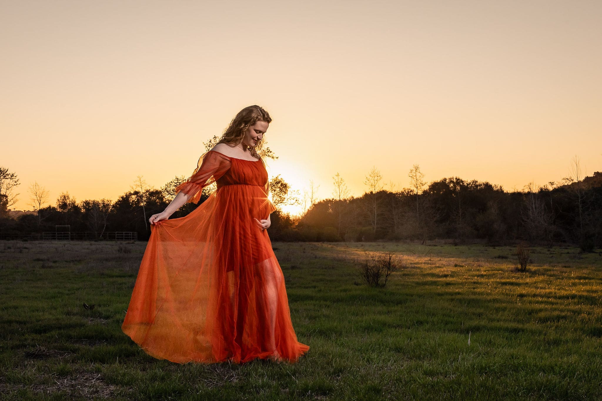 A mom to be in a red maternity gown walks in a meadow at sunset holding her bump and train after meeting doulas in san diego