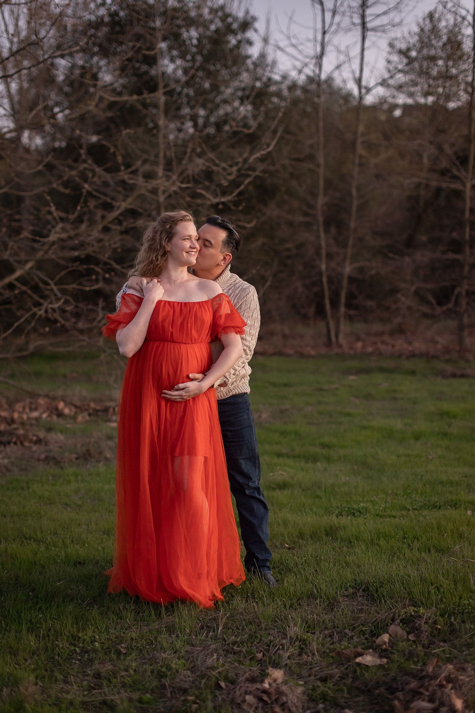 A happy mother to be in a red dress stands in a lawn as he partner kisses her from behind and they hold the bump after meeting doulas in san diego