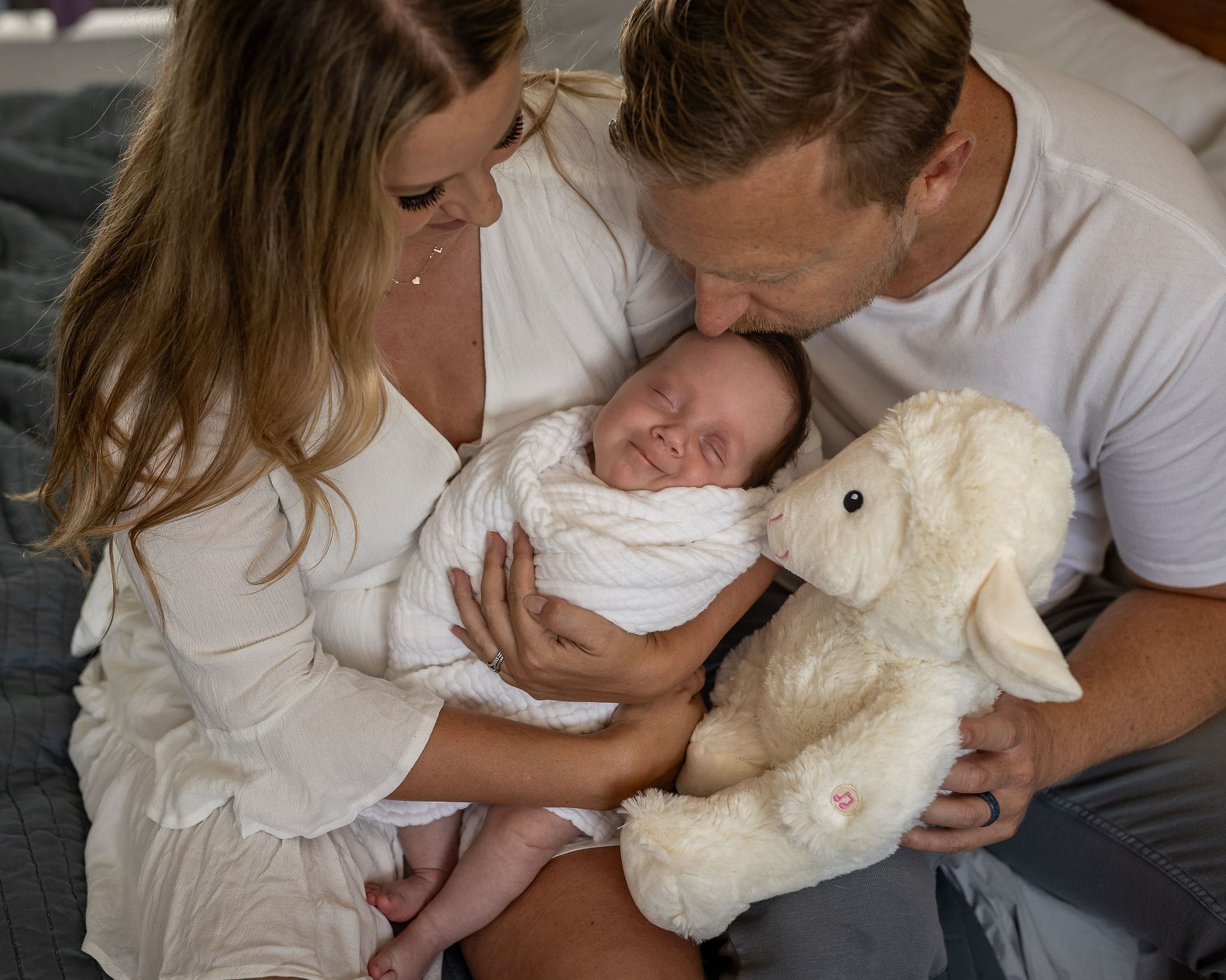 A smiling newborn sleeps in mom and dad's lap with a sheep stuffy as they sit on a bed