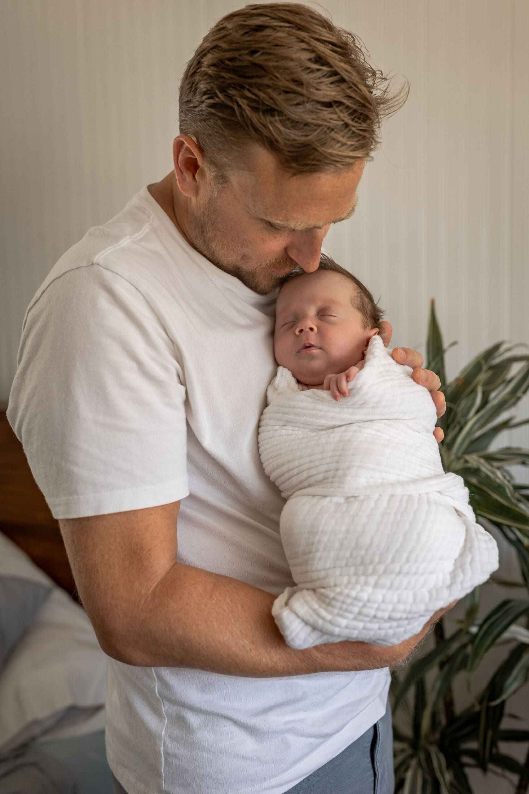 A new dad in white snuggles his sleeping newborn against his chest while standing after meeting lactation consultants in san diego