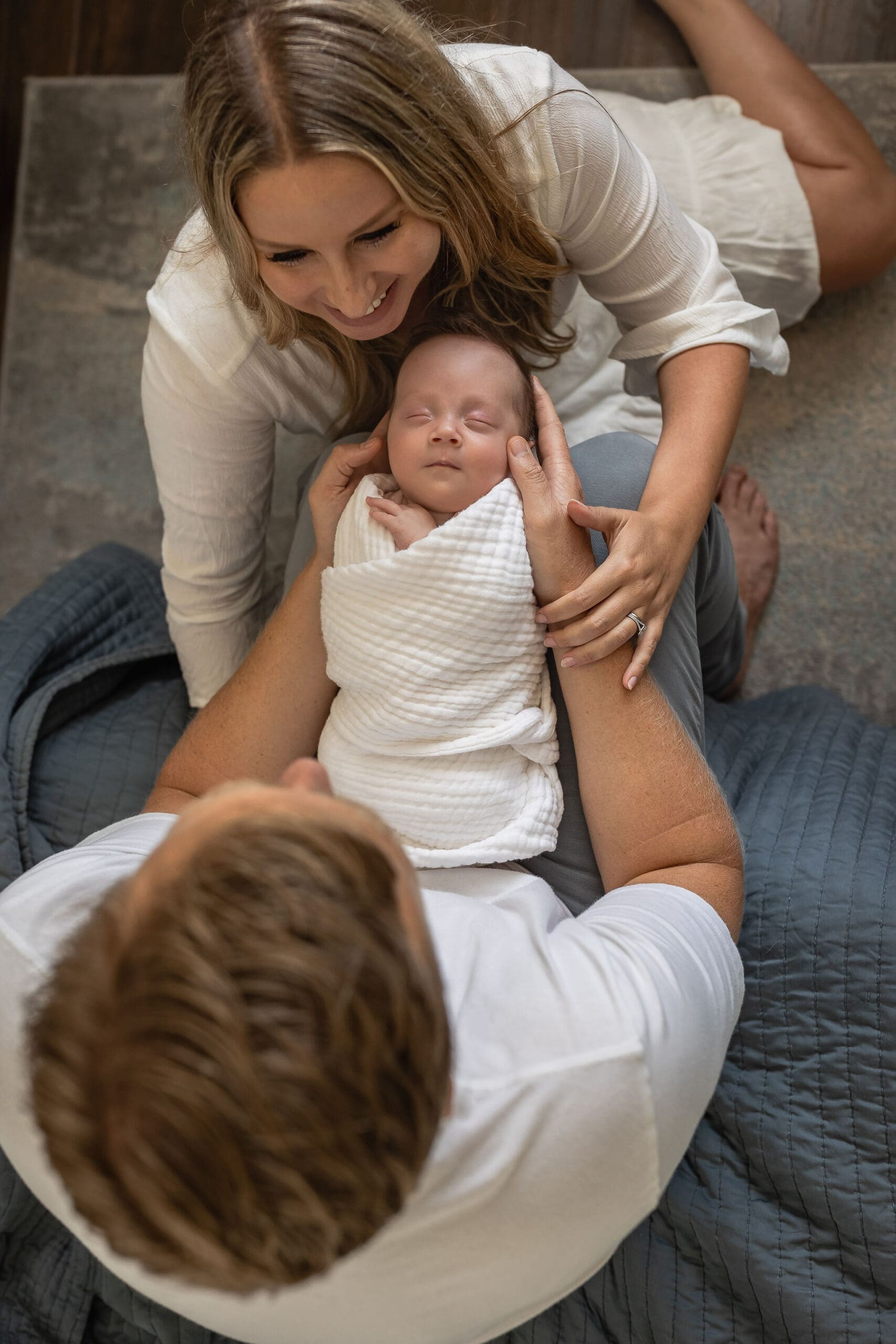 A newborn baby sleeps in a white swaddle in dad's lao as mom leans in from the ground with a smile after meeting lactation consultants in san diego