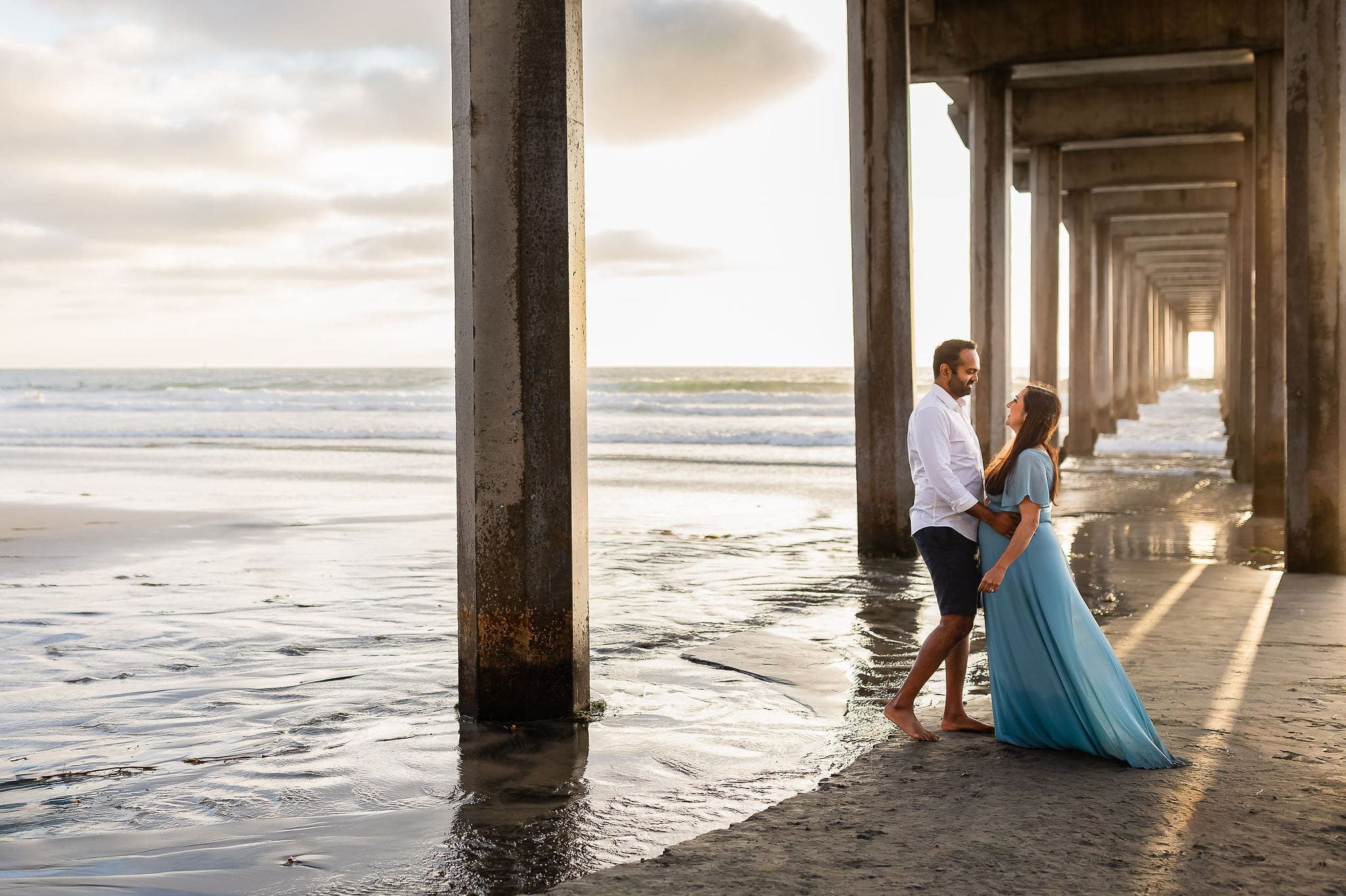 A happy expecting couple cuddle and smile at each other under the beach boardwalk at sunset after meeting midwives in san diego