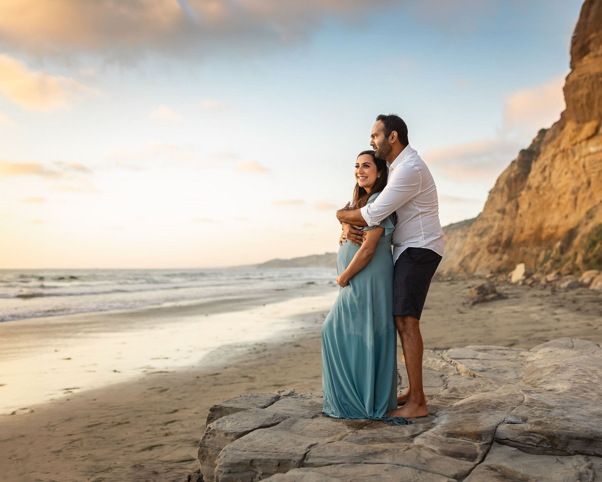 A happy couple embrace at sunset on the beach in a blue maternity gown