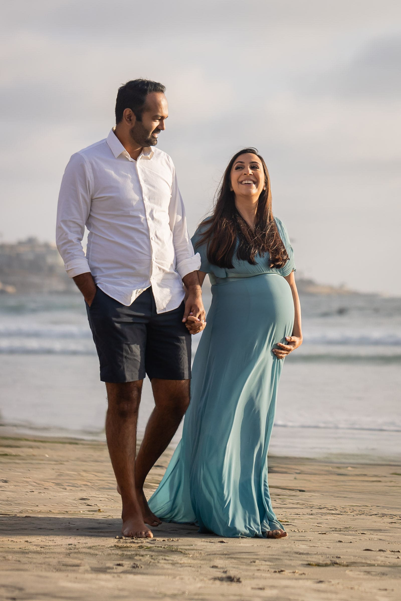 A mother to be in a blue gown walks holding hands with her husband on a beach at sunset laughing after meeting midwives in san diego
