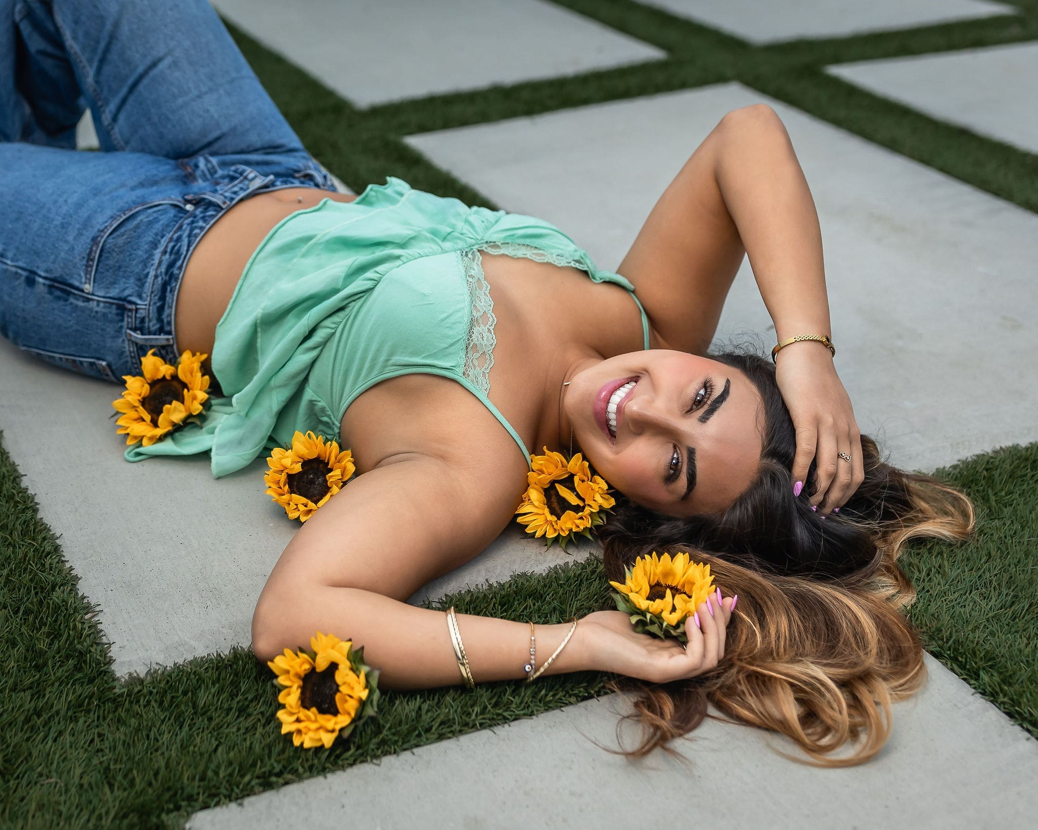 A happy high school grad in a green blouse lays on a patio with sunflowers and big smile and a hand in her hair