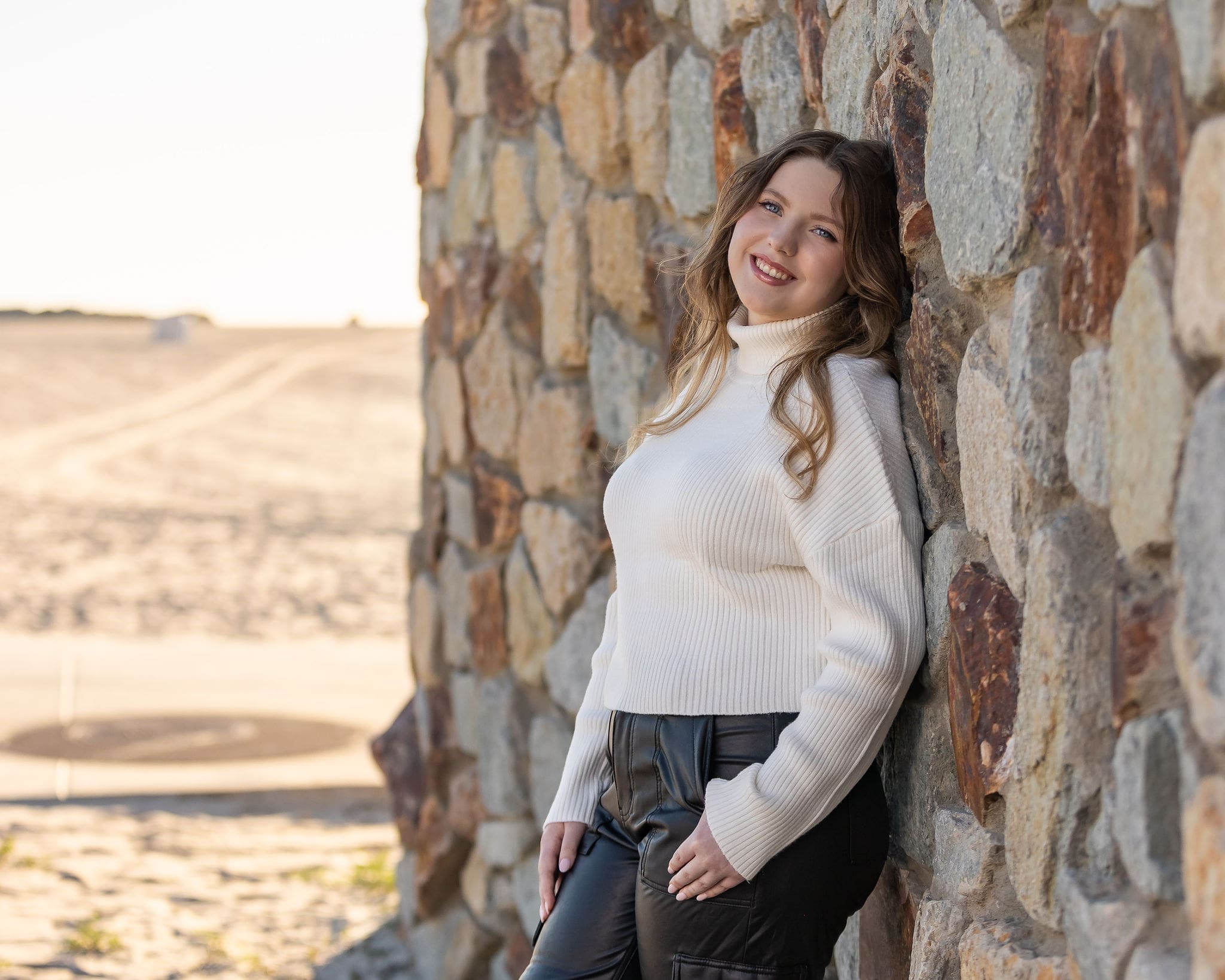A high school senior in a white sweater leans against a stone wall smiling with a thumb in her pocket after visiting nail salons in san diego