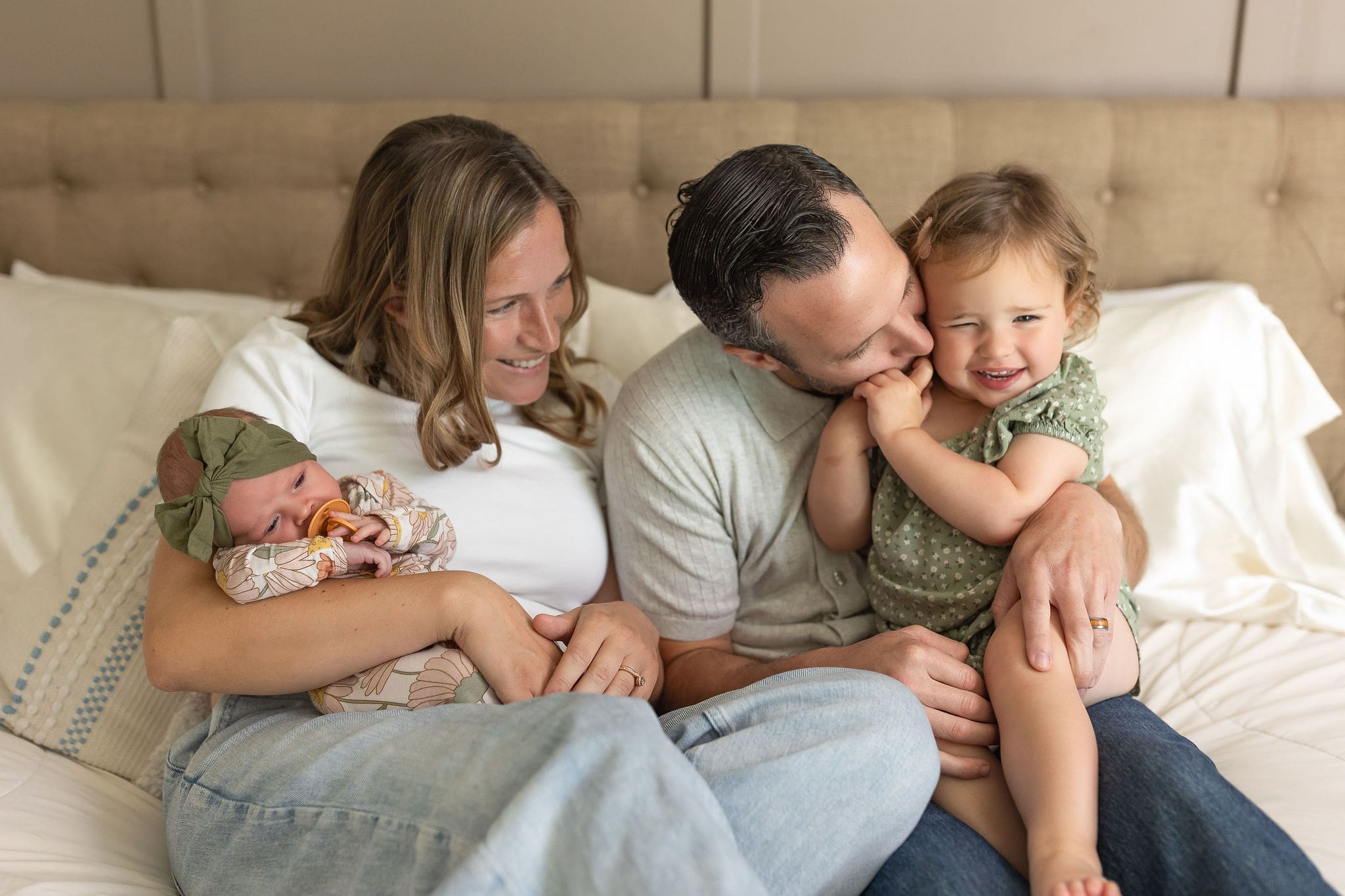 Happy mom and dad cuddle on a couch playing with their toddler daughter and holding a newborn