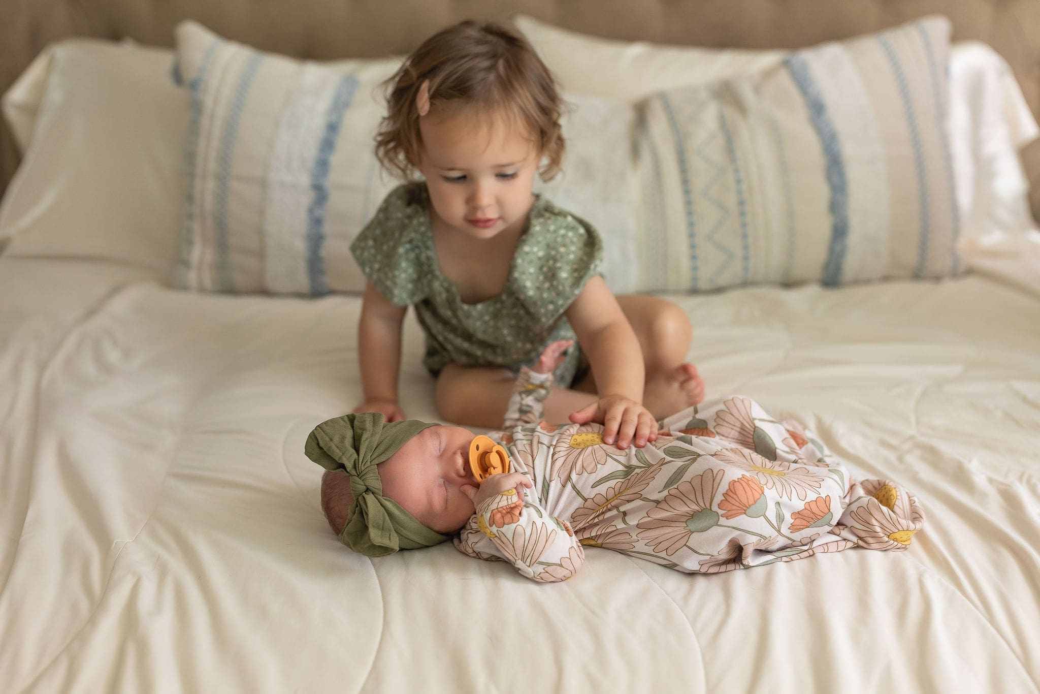 A newborn baby sleeps on a bed in a onesie with big sister reaching to touch her in a green onesie after finding great nannies in san diego
