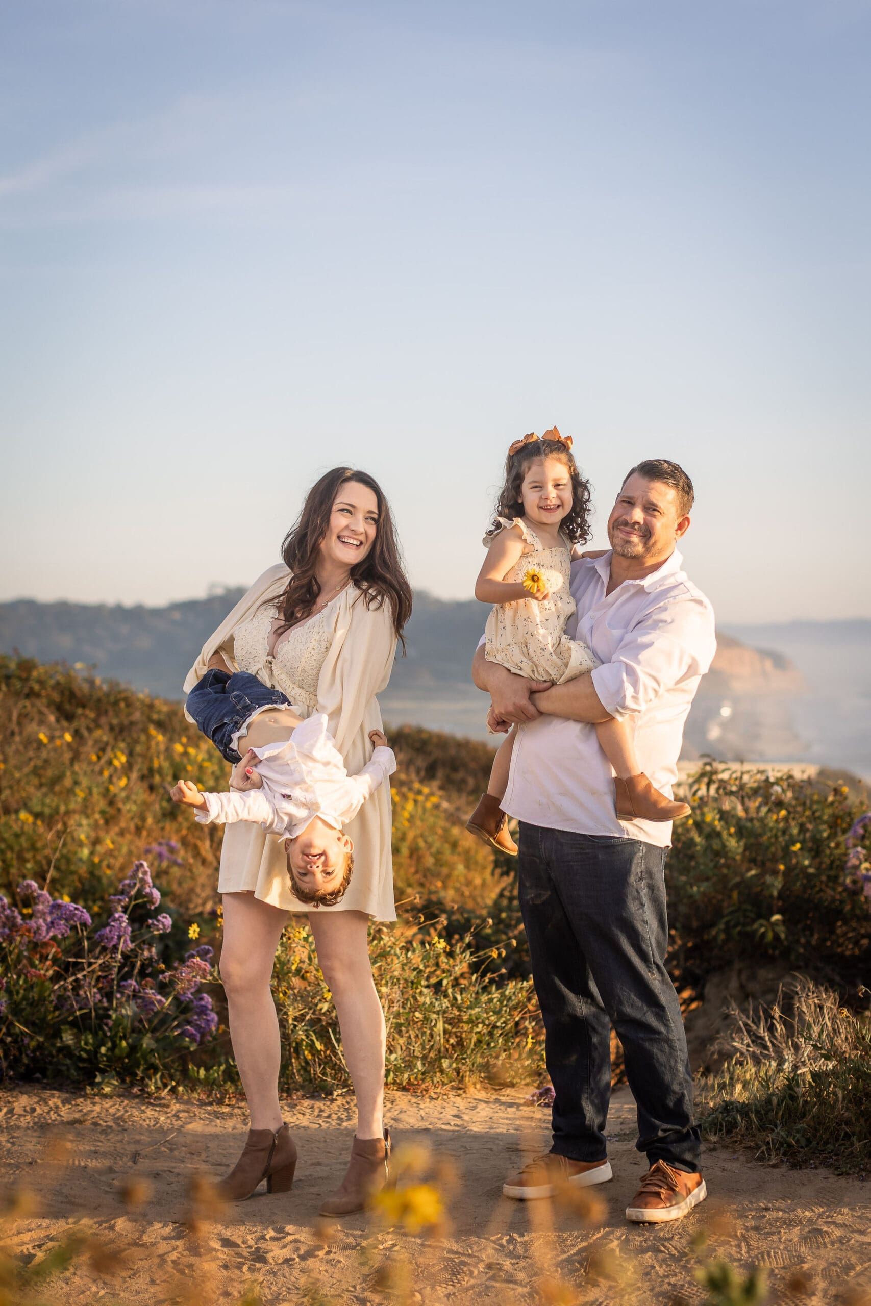 A mother holds and flips over her toddler son while standing on a beach cliff at sunset with dad holding his toddler girl in his arms