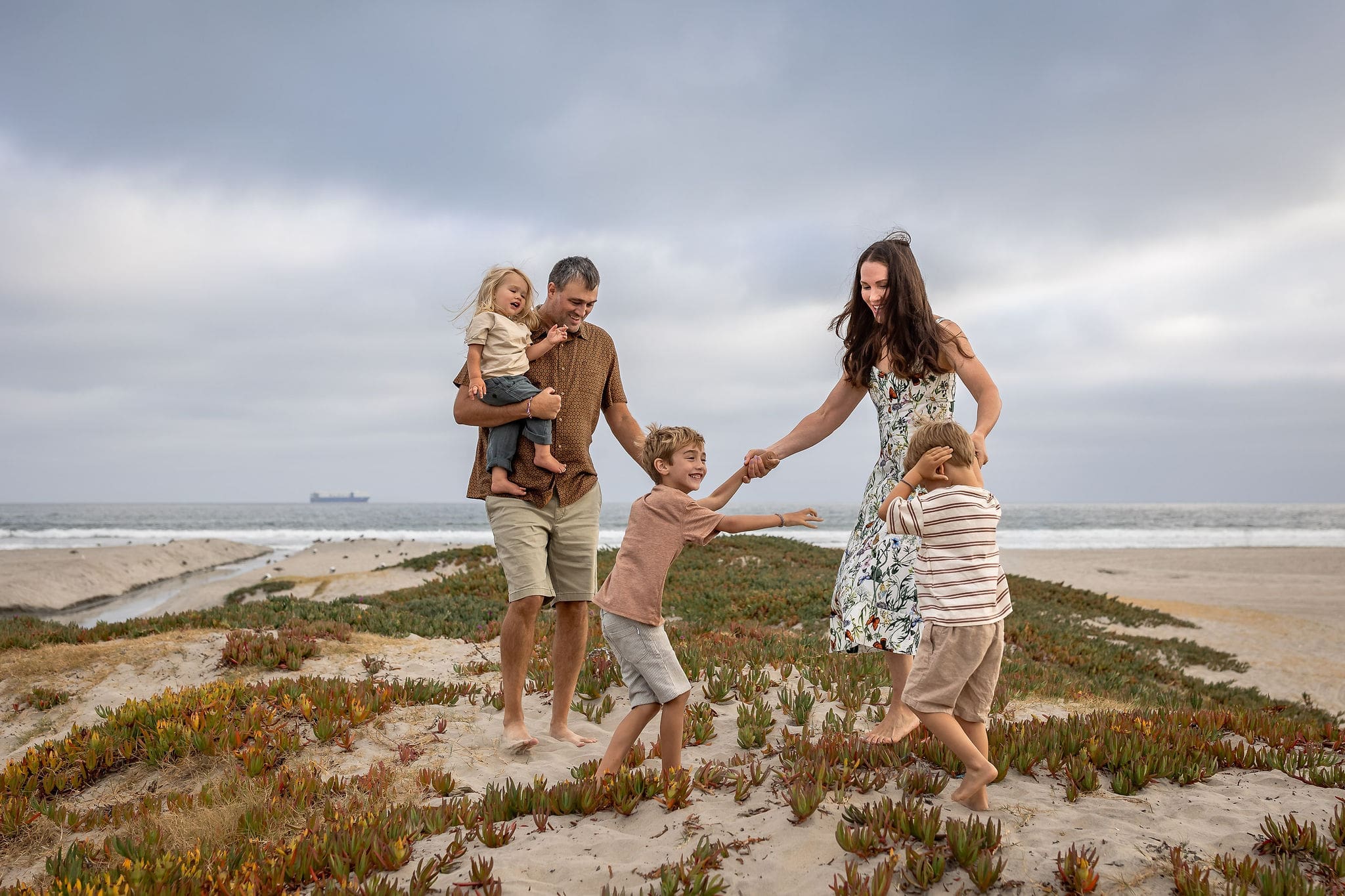 A mom dances with her two toddler sons on a beach dune as dad carries the youngest walking behind them after finding pediatric dentists in san diego