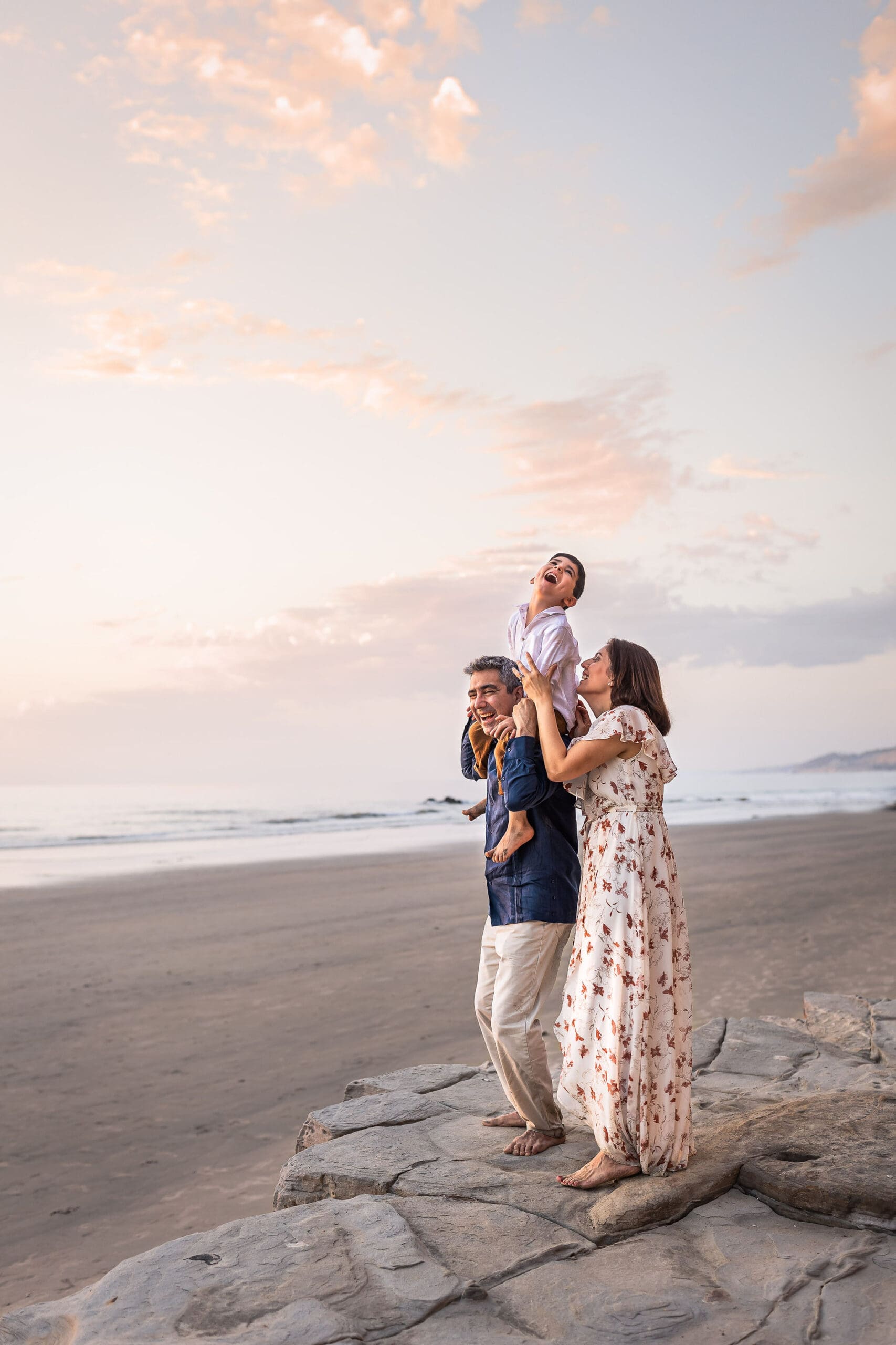 Happy mom and dad play with their giggling son on dad's shoulders on a beach at sunset after finding pediatric dentists in san diego