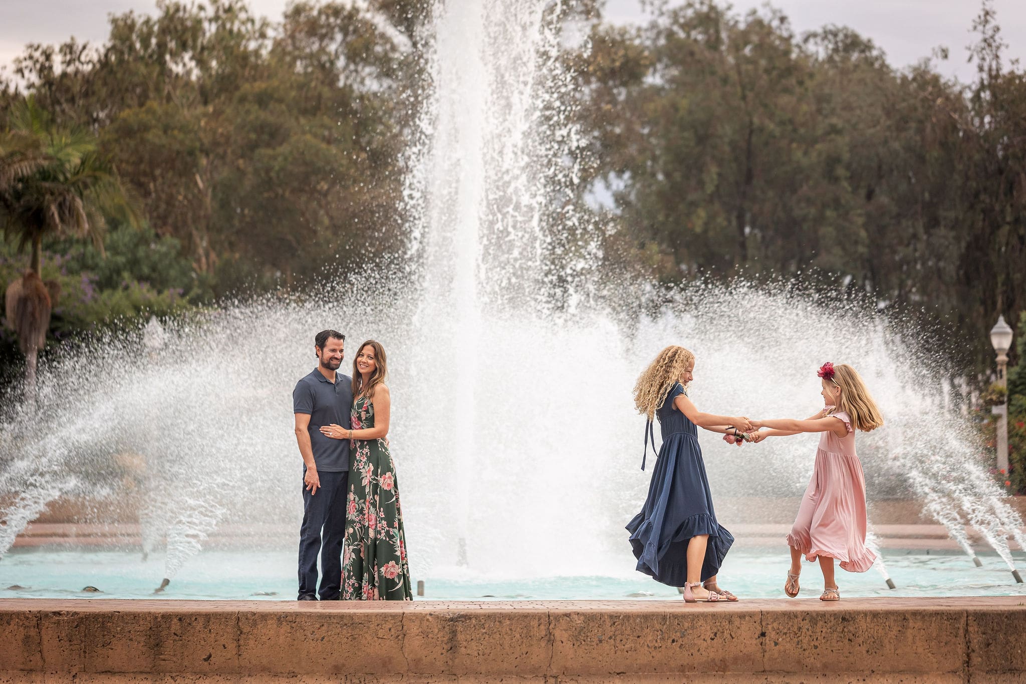 Young sisters in pink and blue dresses dance and spin holding hands while mom and dad watch by a fountain in a garden