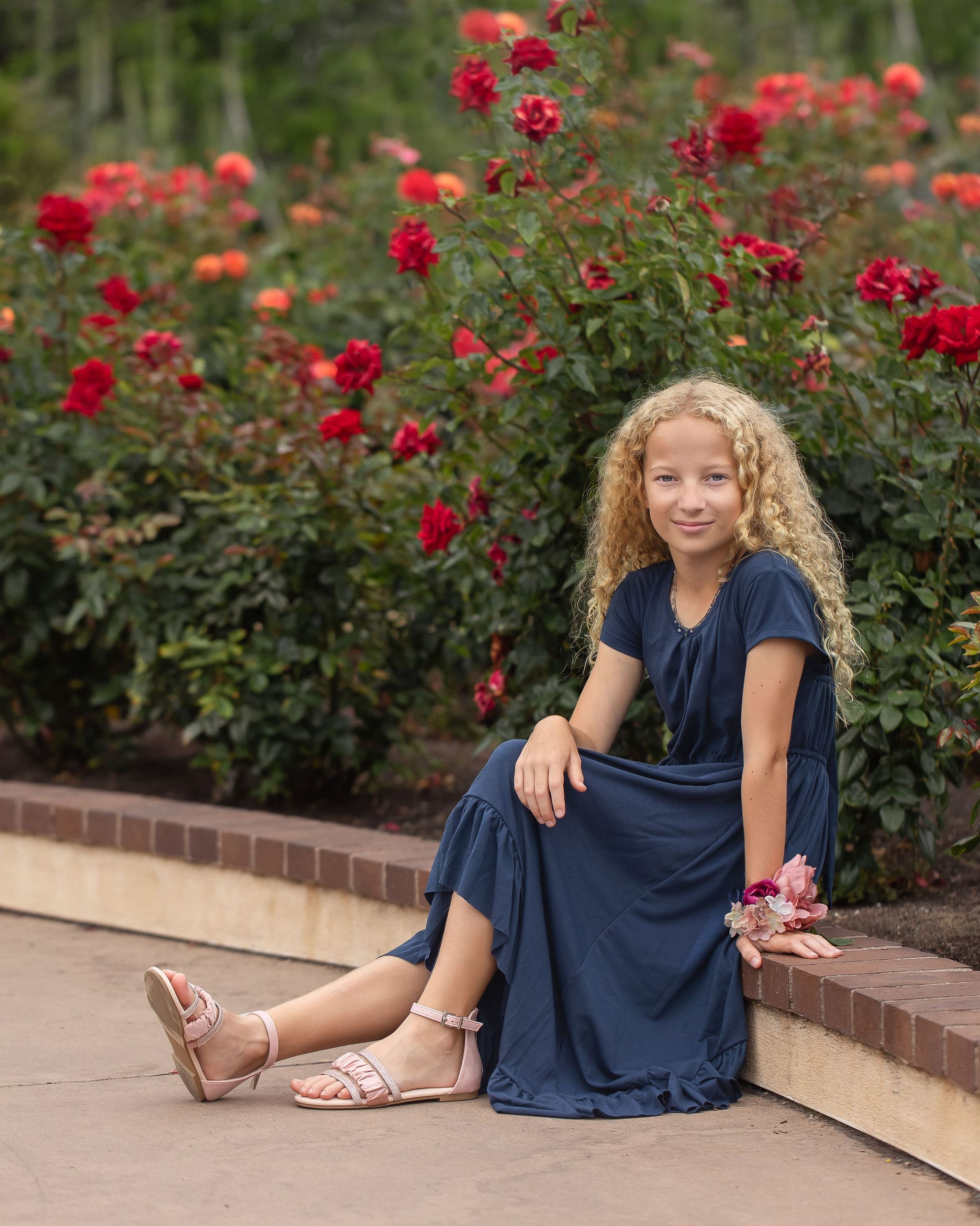 A young girl in a blue dress sits on on the edge of a rose garden smiling after visiting pediatricians in san diego