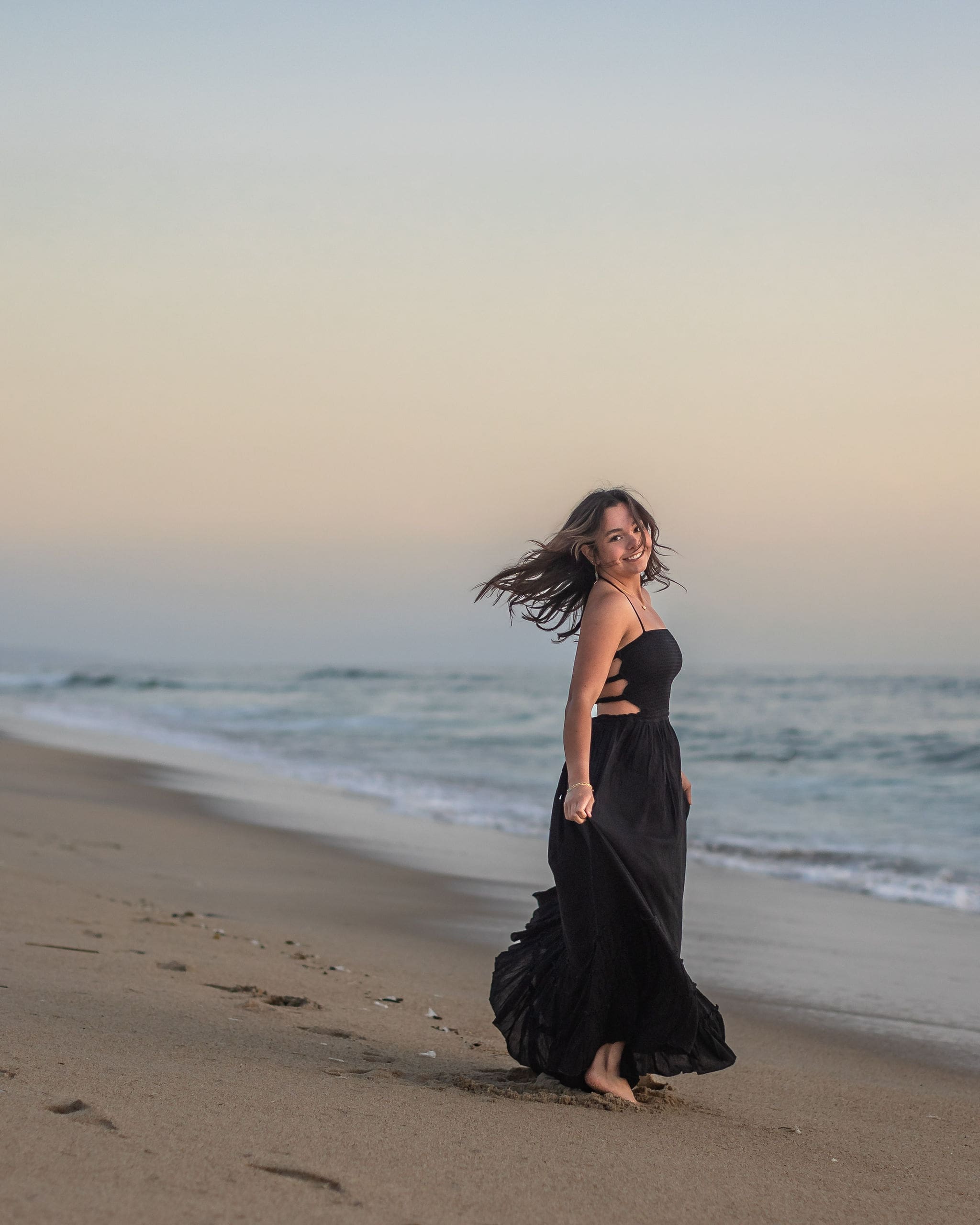 A dancing high school senior in a black dress spins around while playing on a beach at sunset after finding prom dresses in san diego