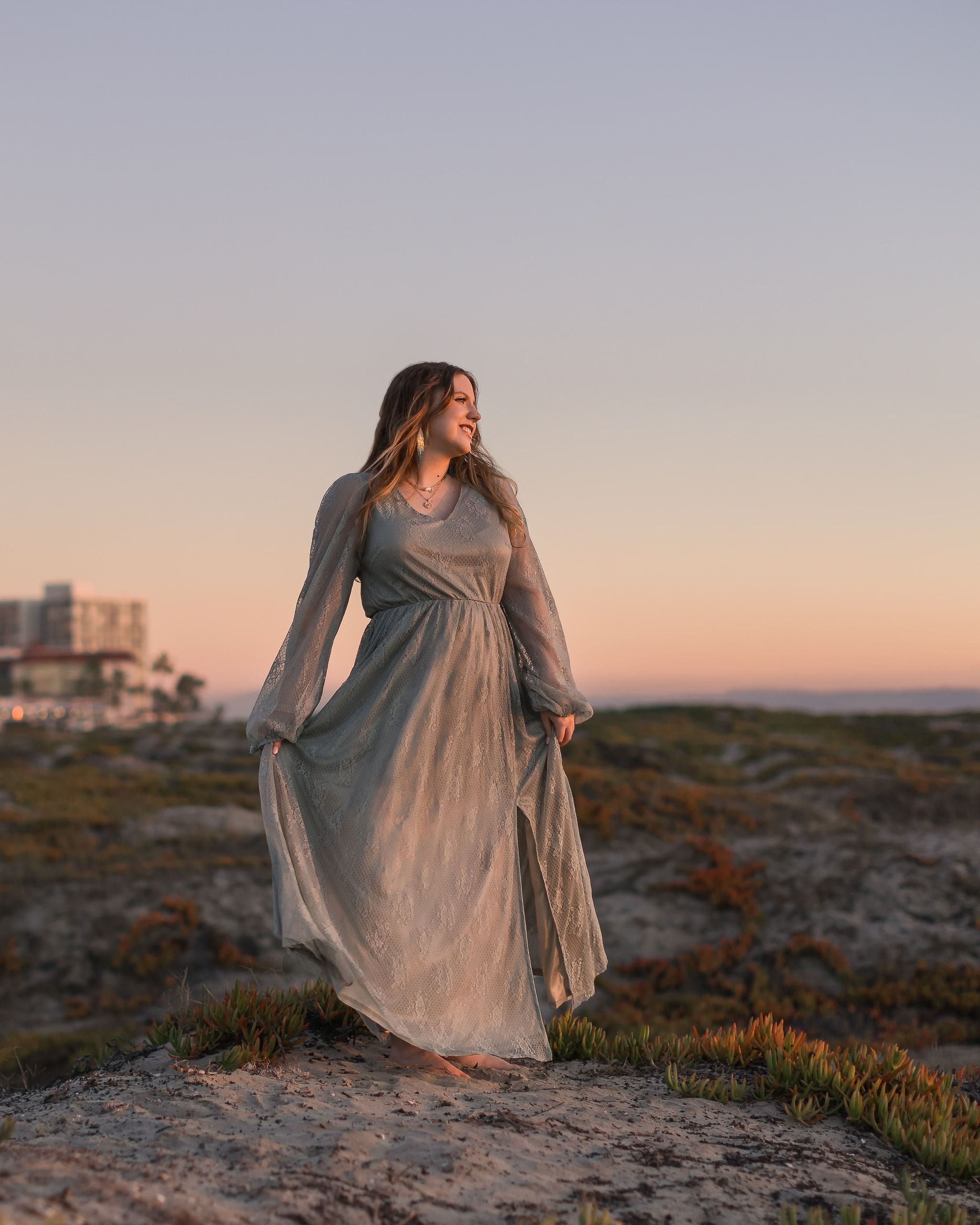 A happy high school senior in a long gown stands smiling in the sunset glow on a beach after finding perfect prom dresses in san diego