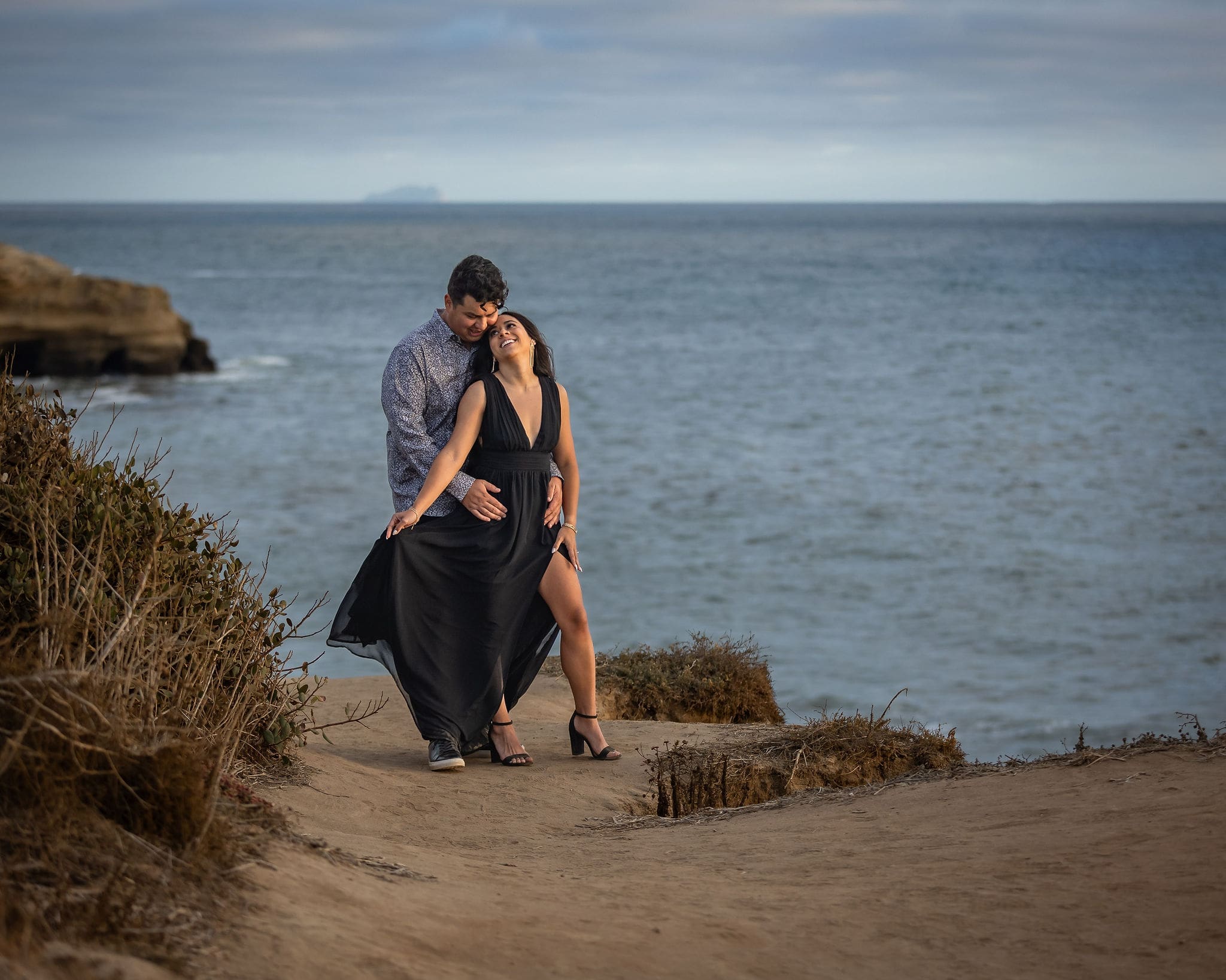 A woman in a black dress leans into her fiancee on a beach overlook at sunset as they cuddle