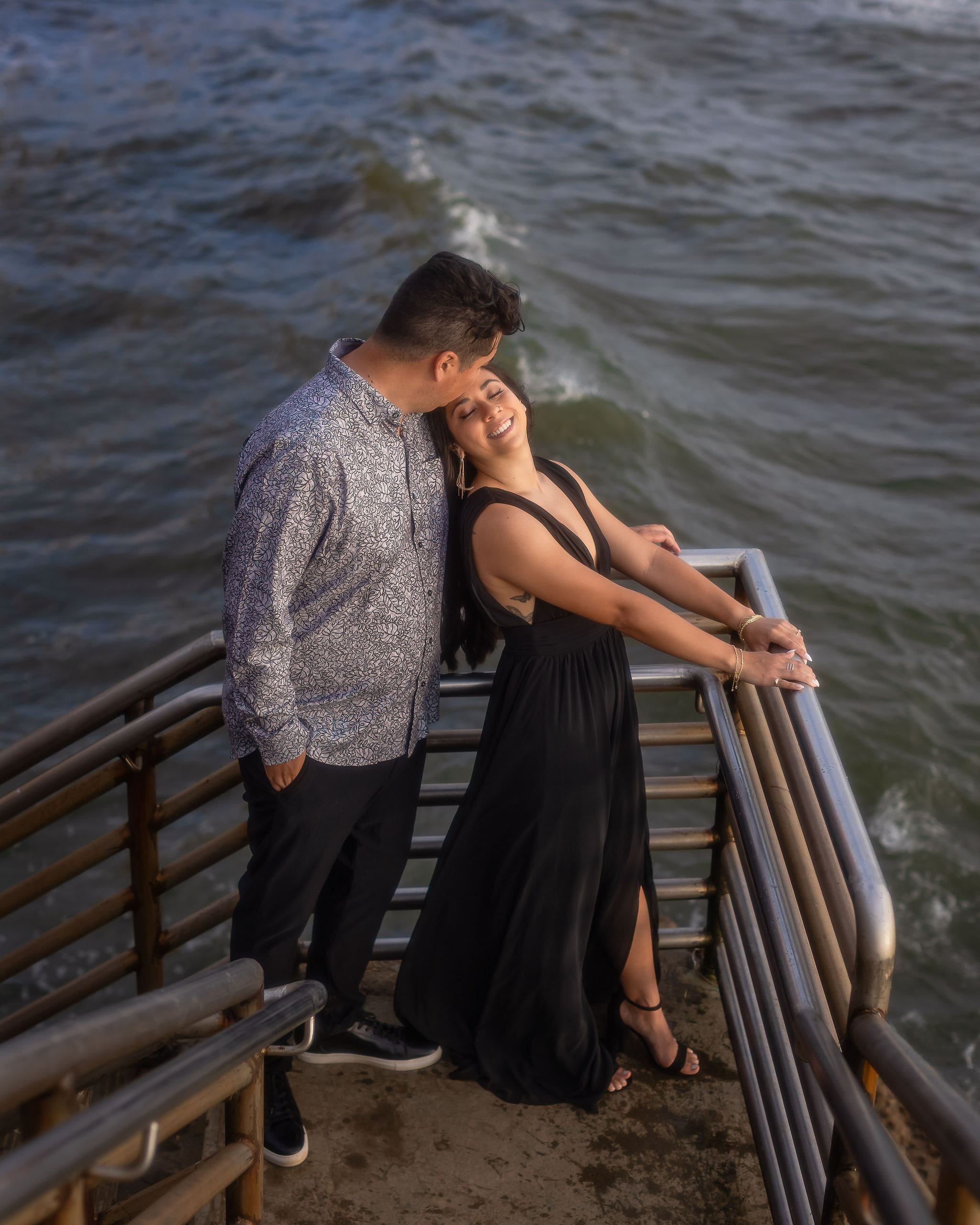 A happy couple in black and grey stands on an ocean stairwell enjoying the sunset after finding romantic restaurants in san diego
