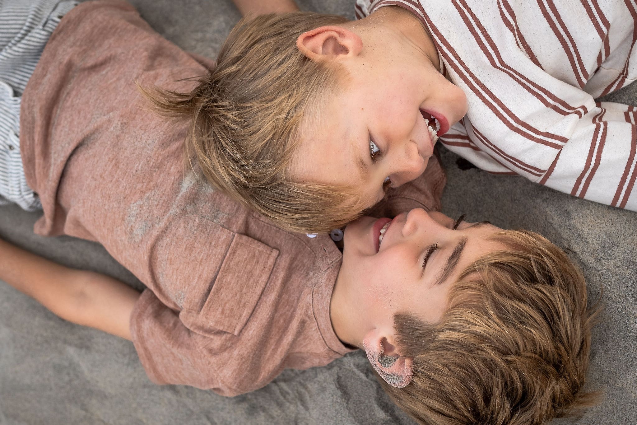 Toddler brothers lay face to face on the beach giggling before visiting toy stores in san diego