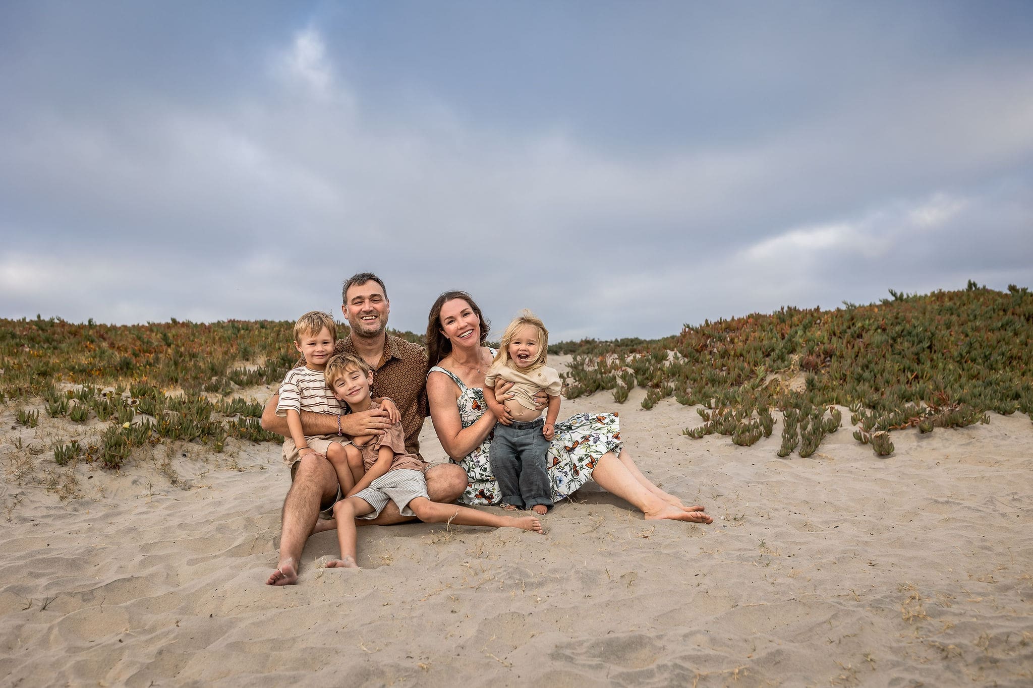 Happy mom and dad sit on a beach laughing with their three toddlers in their laps before visiting toy stores in san diego