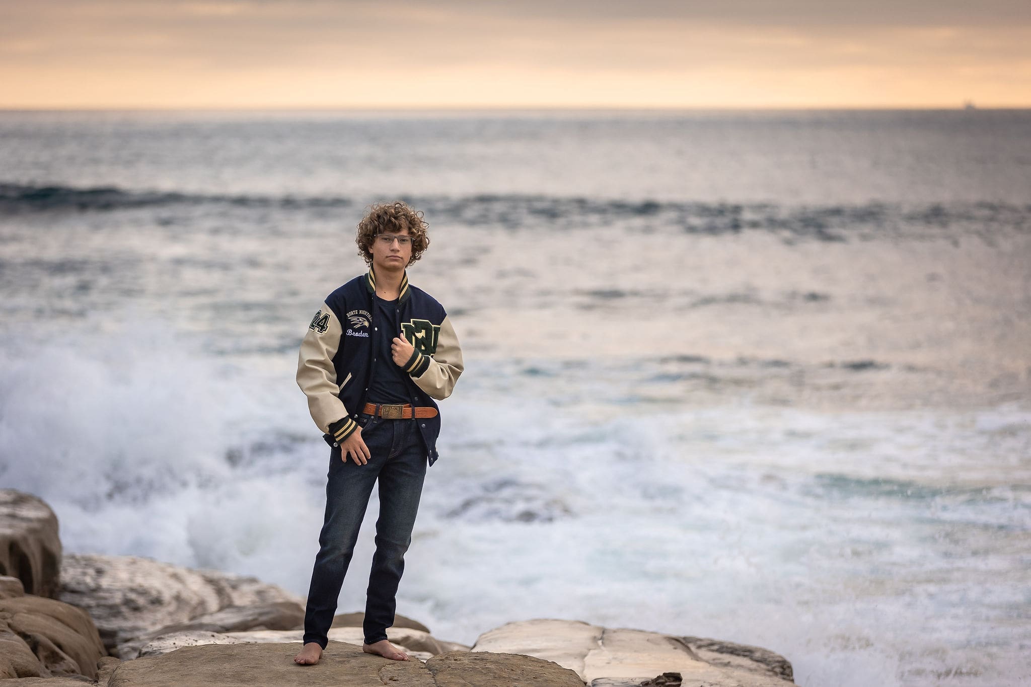 A high school grad stands on a boulder on the beach in his letterman jacket at sunset