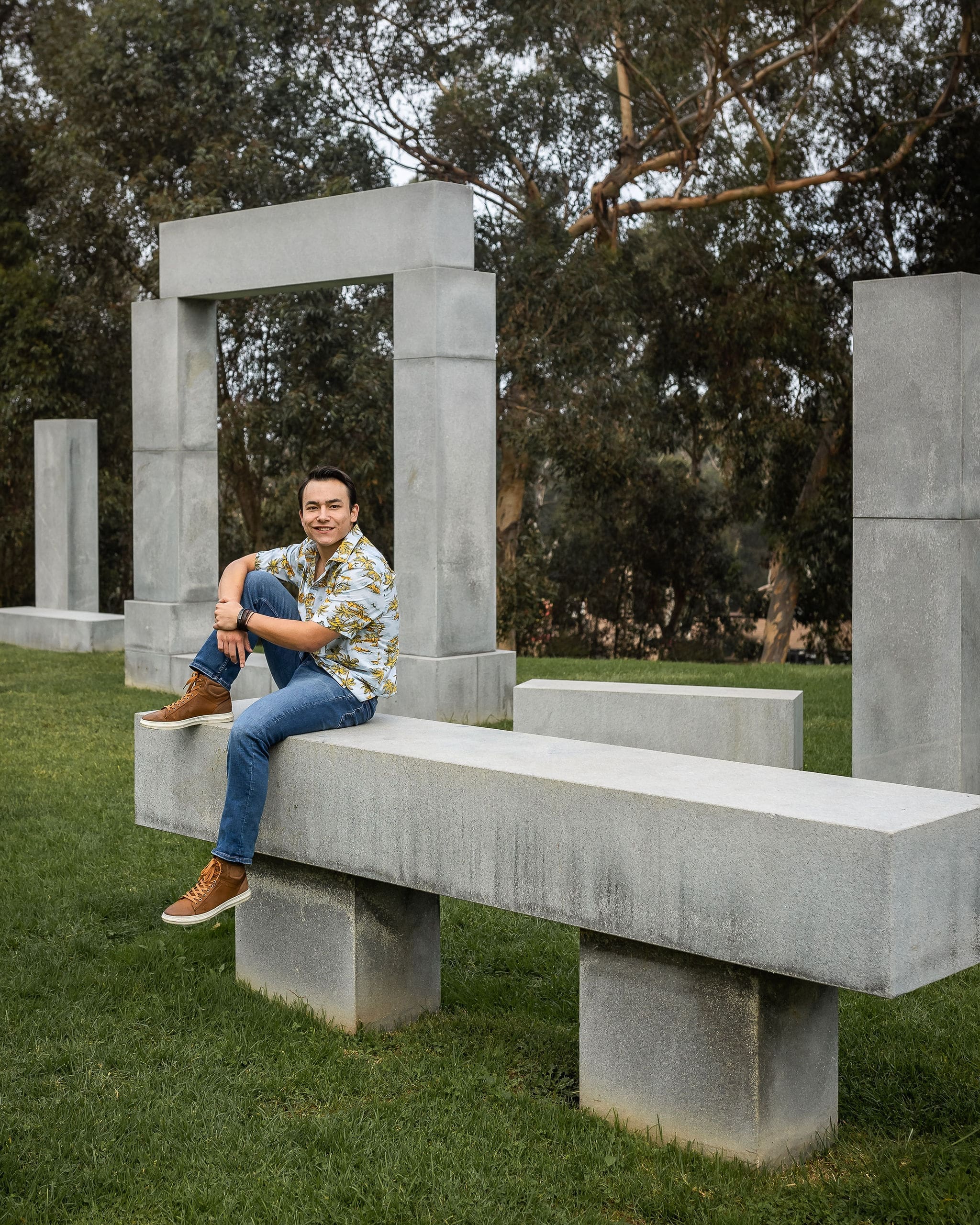 A happy high school senior sits on a concrete sculpture in a park in a colorful shirt and jeans after meeting college counselors in san diego