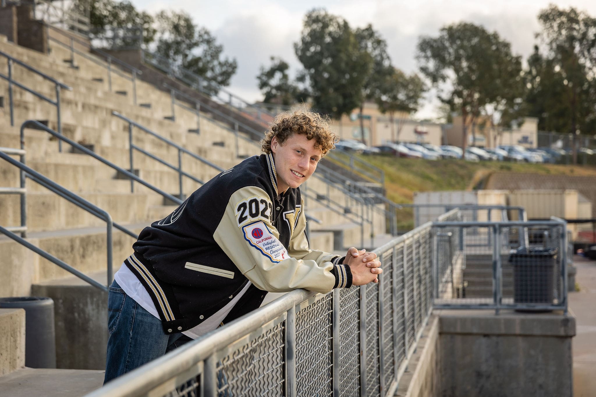 A high school senior leans against a fence railing in a football stadium at sunset in his letterman jacket after meeting college counselors in san diego