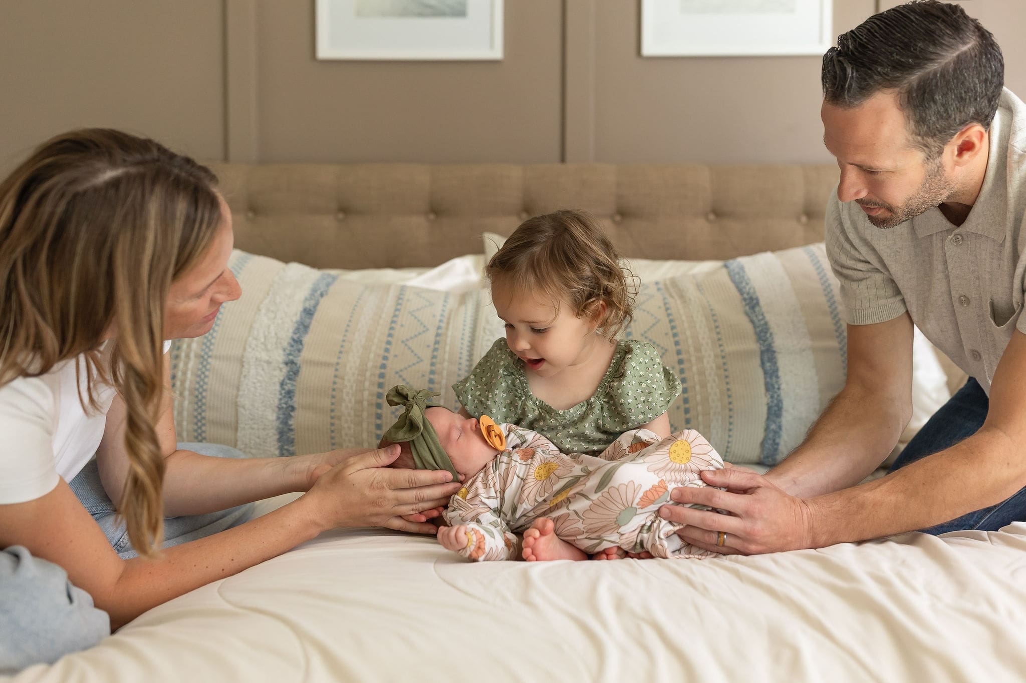 A toddler girl in a green dress meets her newborn sister in a bed with mom and dad's help while using a diaper service in san diego