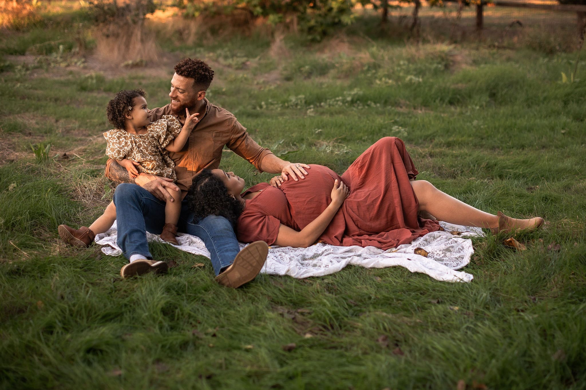 A dad rests his hand on wife's pregnant bump while she lays in the grass resting her head on his leg and he chats with toddler in his lap