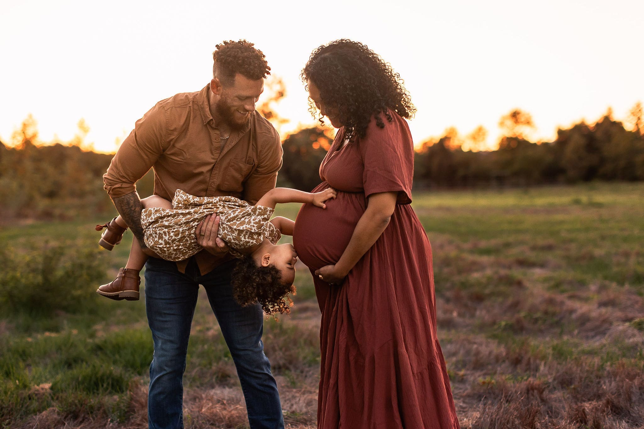 An upside down girl kissing mom's pregnant belly at sunset in a park while being held by dad after mom had a prenatal massage in san diego