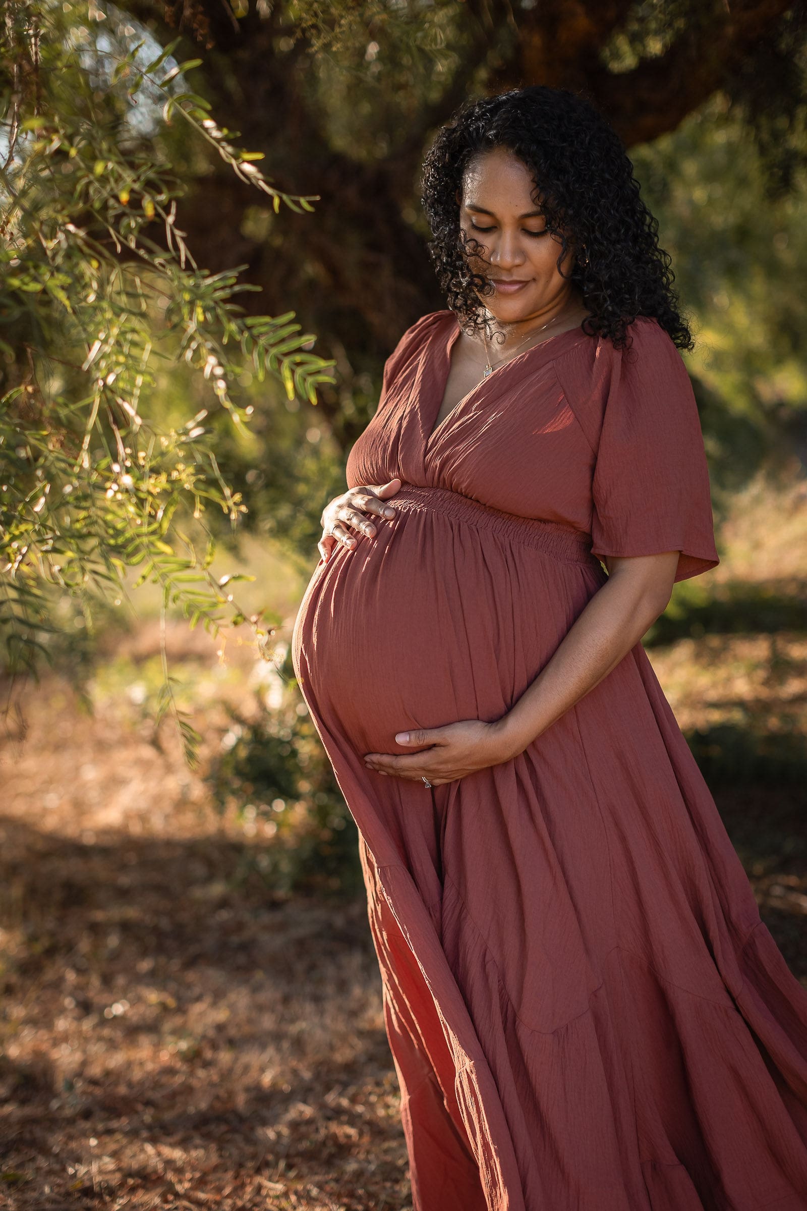 A mother to be in a mauve maternity gown stands under a tree at sunset holding her bump after a prenatal massage in san diego