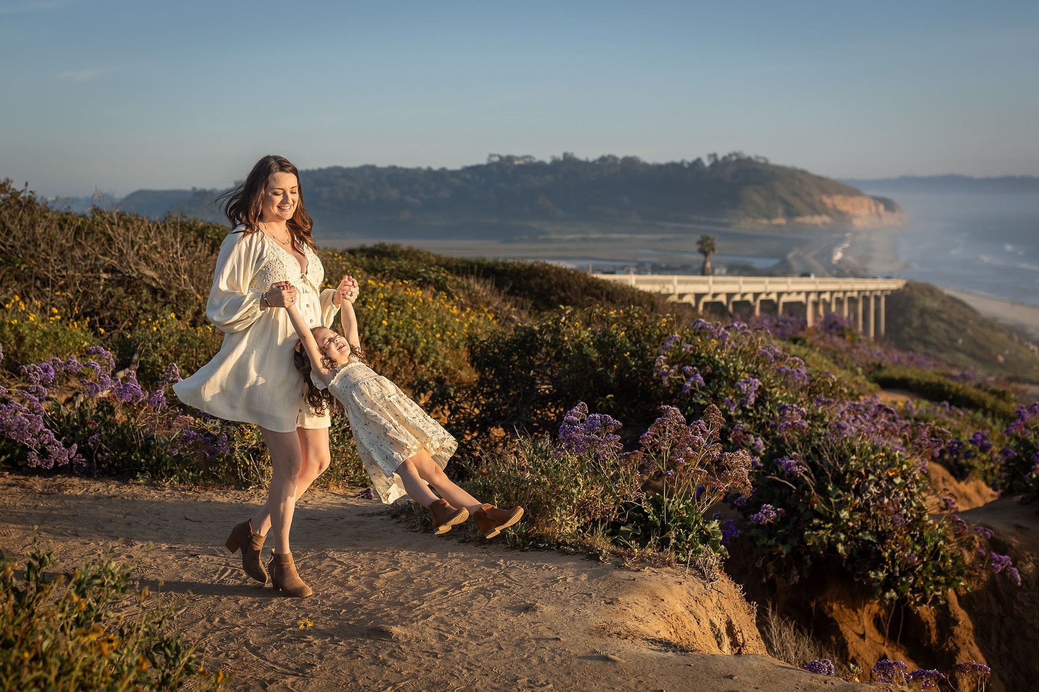 A mom spins her toddler daughter in matching white dresses on an overlook at sunset