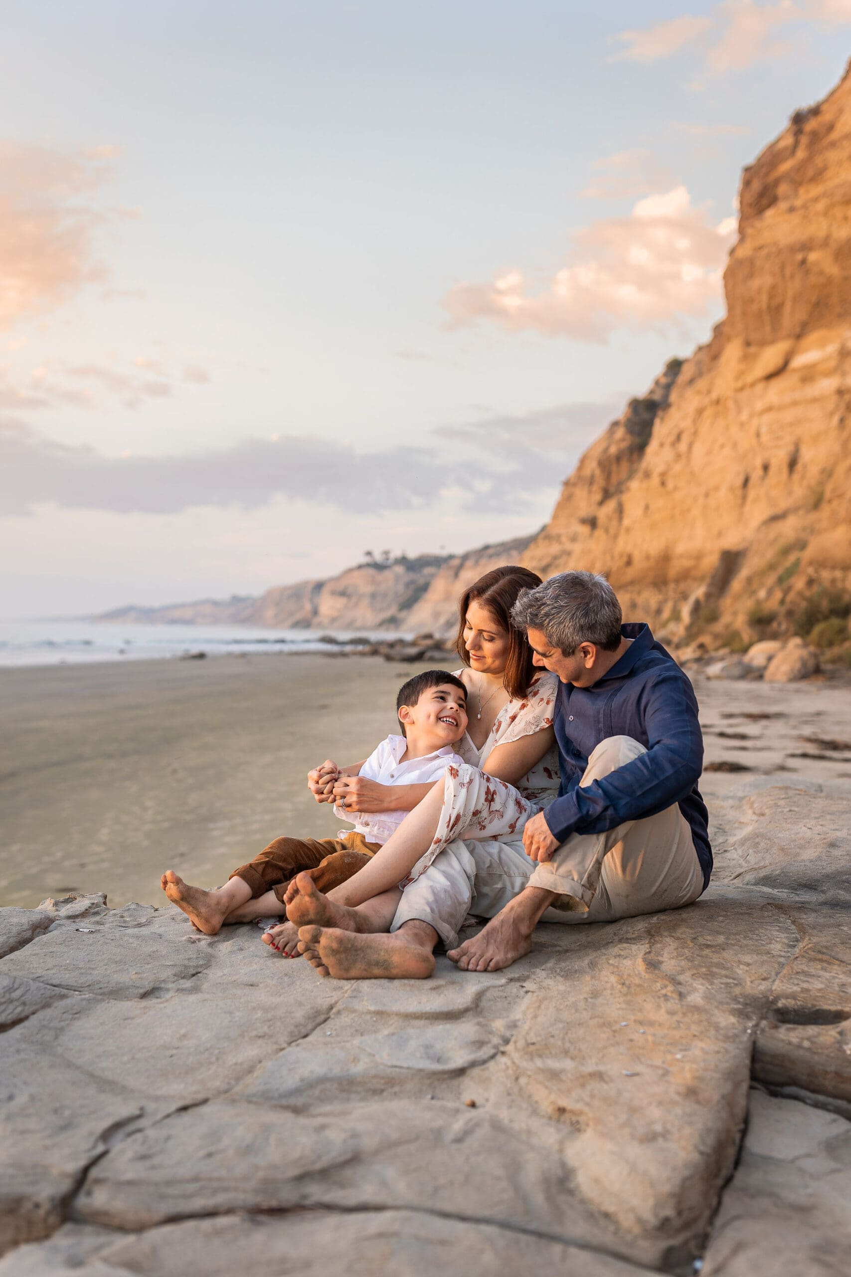 A happy toddler boy in white sits in mom's lap playing on a beach rock with dad after enjoying preschools in san diego