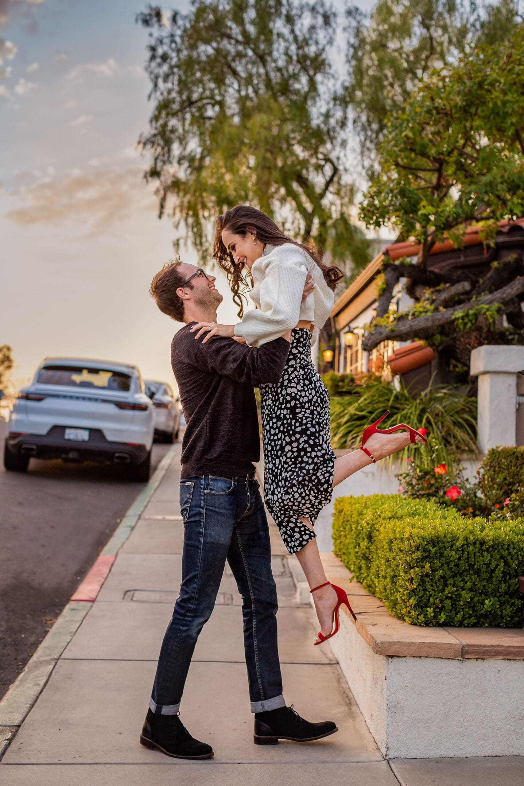 A man in a dark sweater lifts his fiancee while walking on a sidewalk at sunset after staying at romantic hotels in san diego